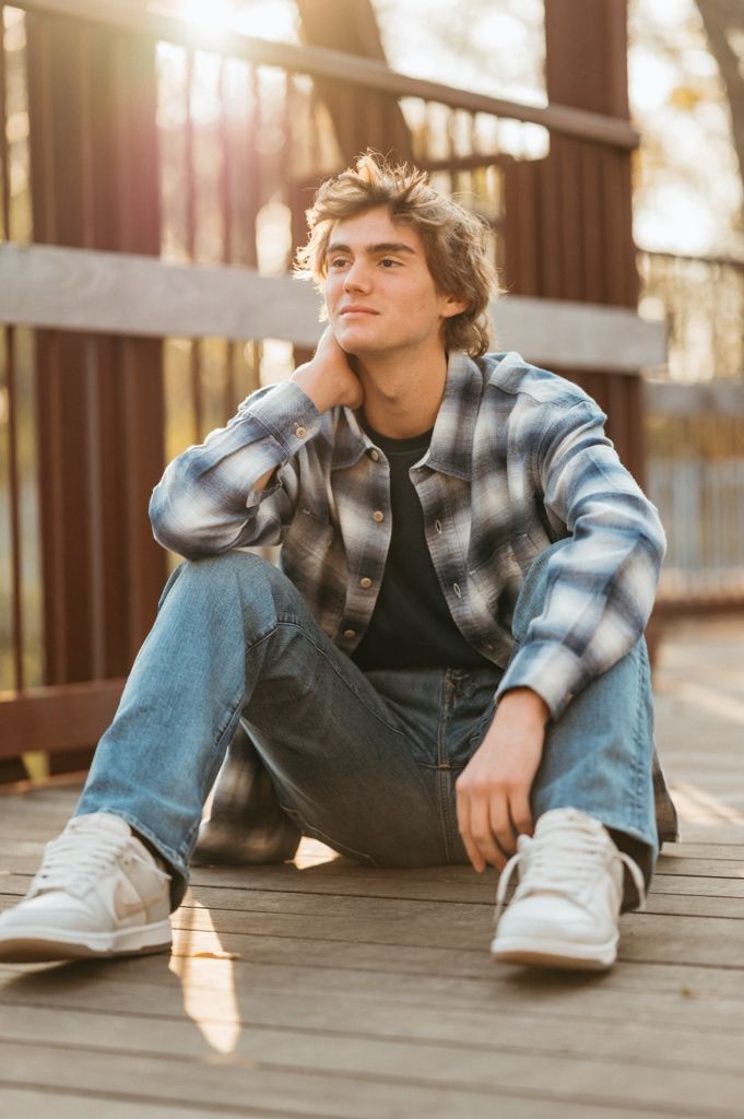 Person in plaid shirt and jeans sits casually on wooden deck in warm sunlight.