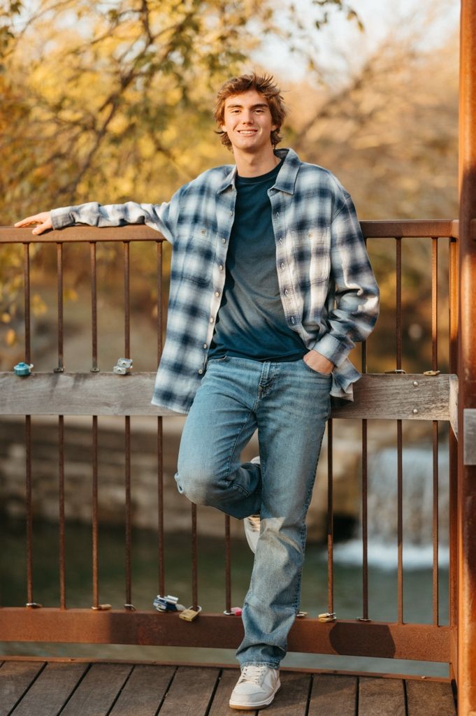 Person in plaid shirt and jeans poses casually against rustic railing during golden hour photoshoot.