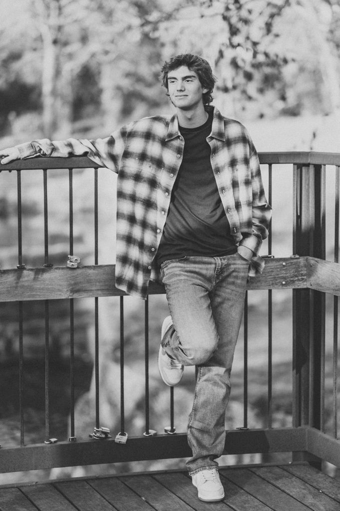 Black and white portrait of someone in a plaid flannel shirt and jeans leaning casually against a wooden deck railing.
