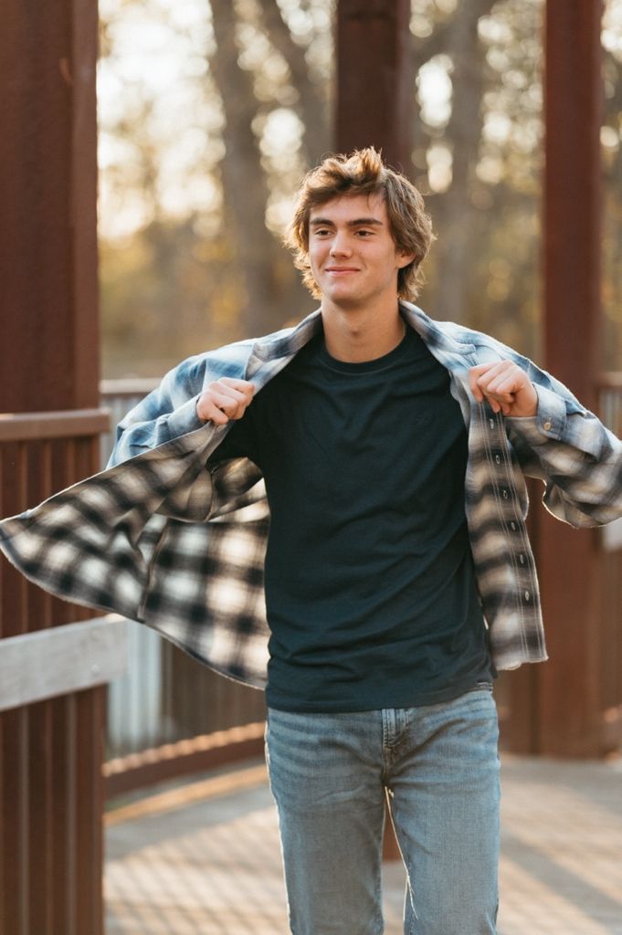 A person in a black sweater and plaid flannel shirt poses playfully against wooden railings at sunset.