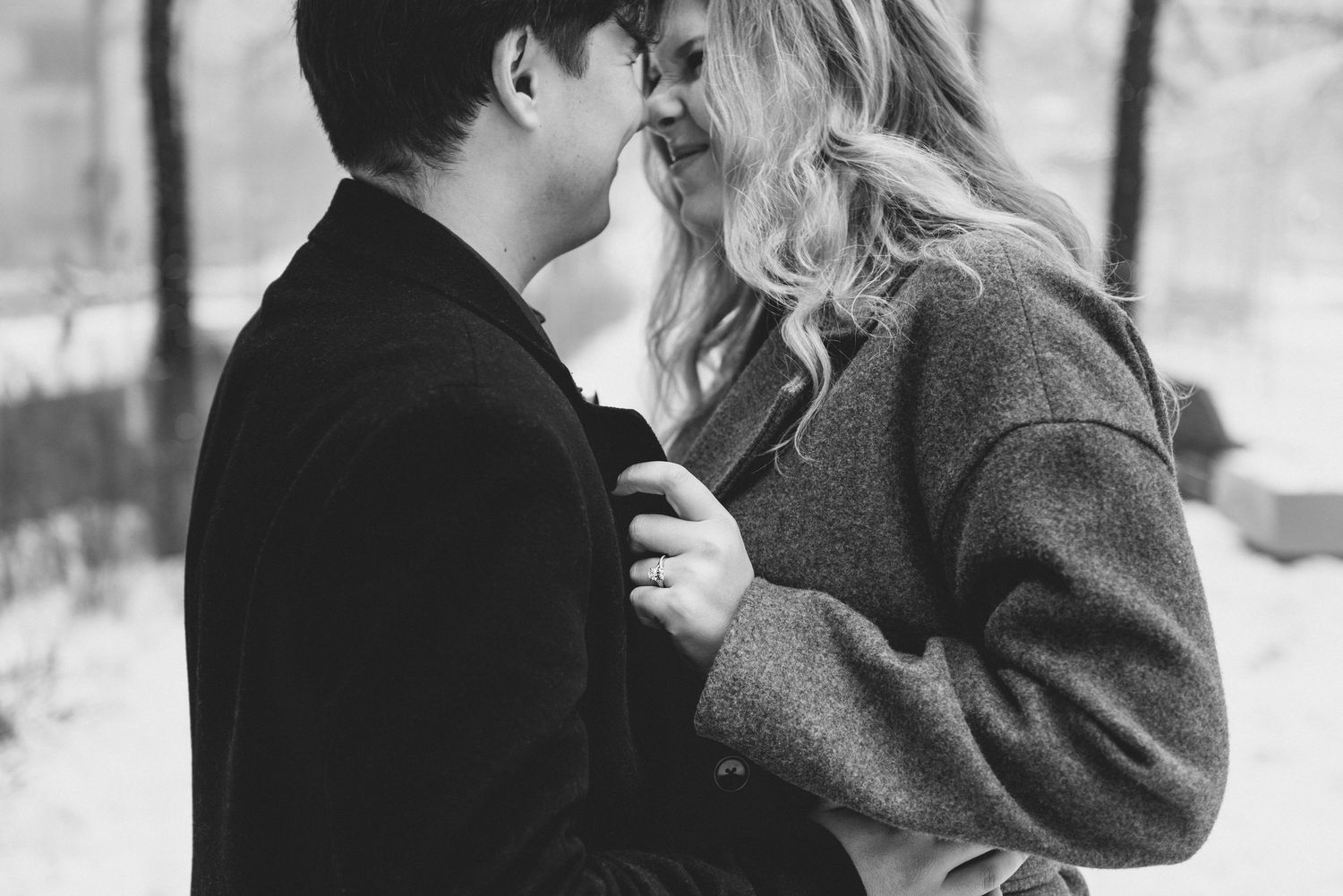 A couple shares a romantic embrace in a snowy winter scene wearing cozy coats and scarves.