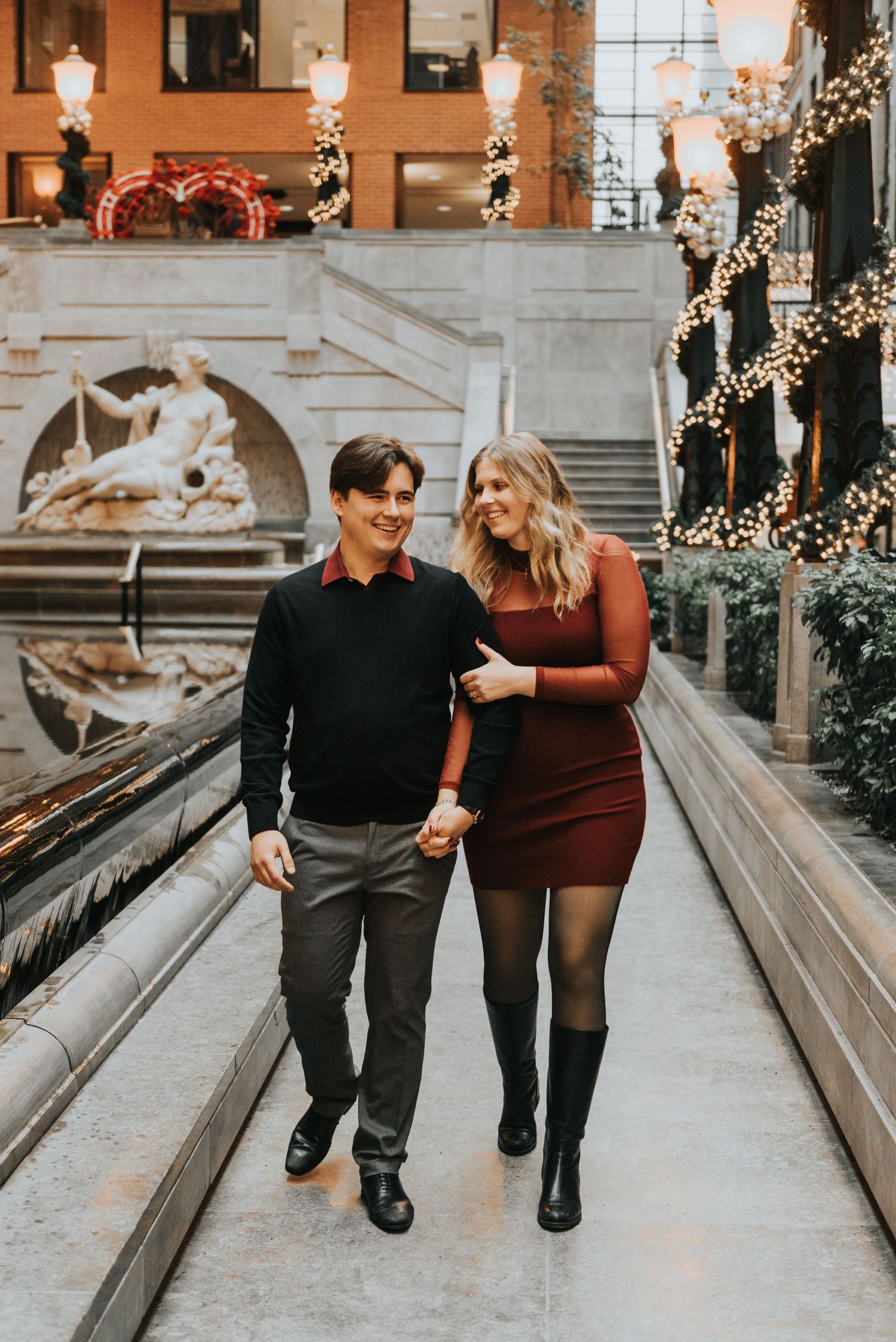 A couple walks together on a brick-lined path decorated with twinkling holiday lights and festive winter decor.