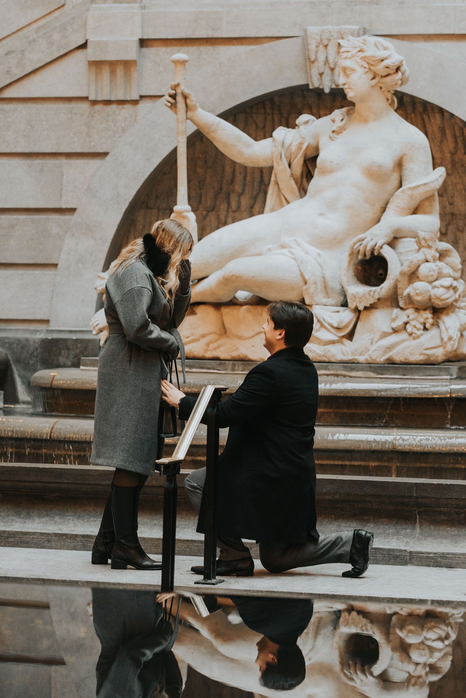 A romantic proposal sequence showing a couple by a classical fountain as one partner gets down on one knee.