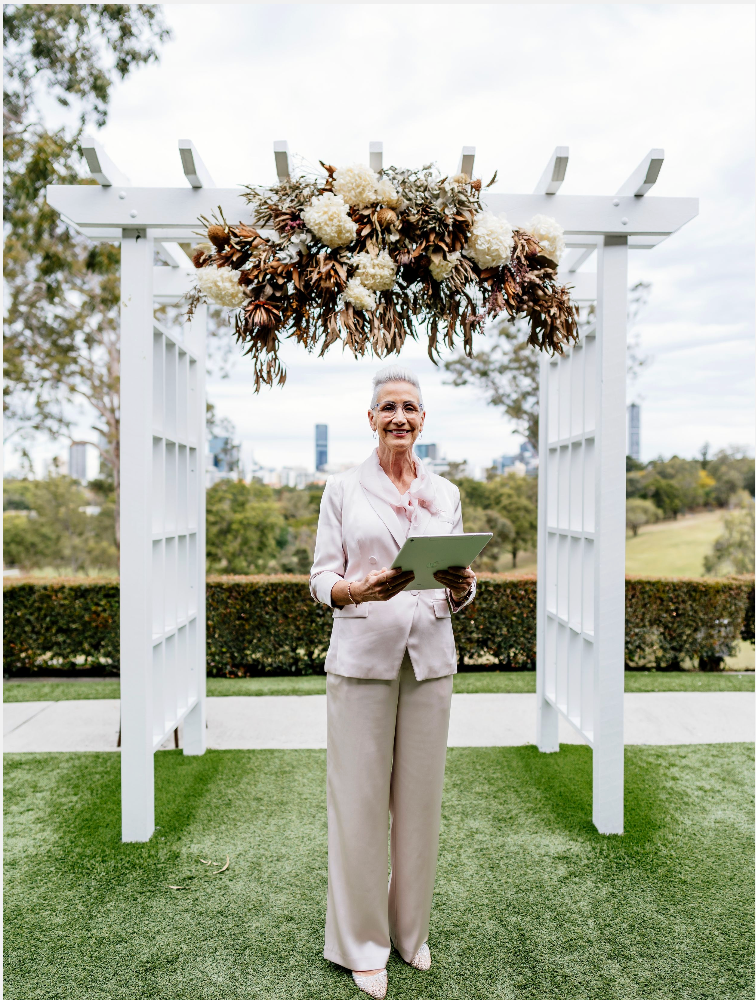 A woman in a white floral dress with flutter sleeves poses outdoors with greenery and city buildings in the background.