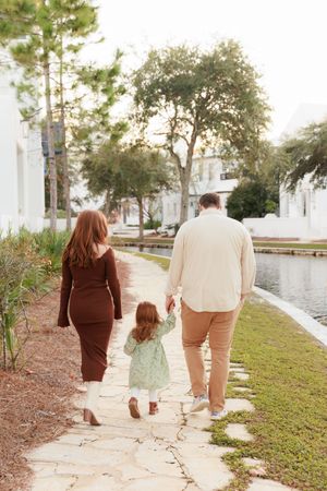 A couple walks together on a stone path near a scenic waterfront surrounded by lush greenery and trees.