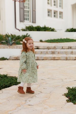 A small child in a green floral dress and brown boots walks on stone pavement near a white building.