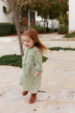 A young child wearing a mint floral dress and brown boots spins playfully on a stone patio in an outdoor setting.