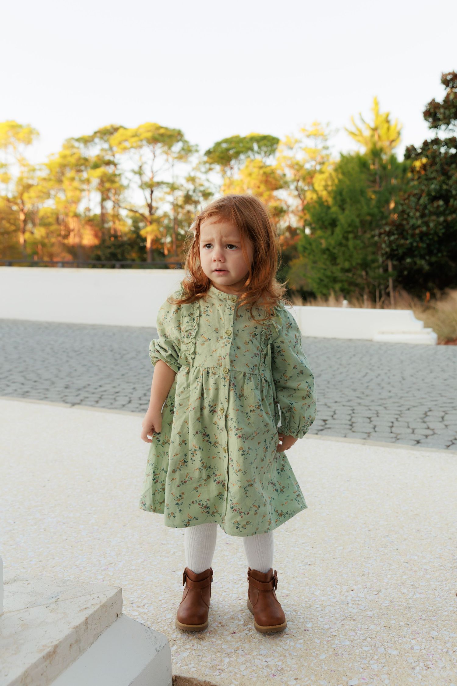 A small child wearing a floral green dress and brown boots plays on a sandy beach at sunset.