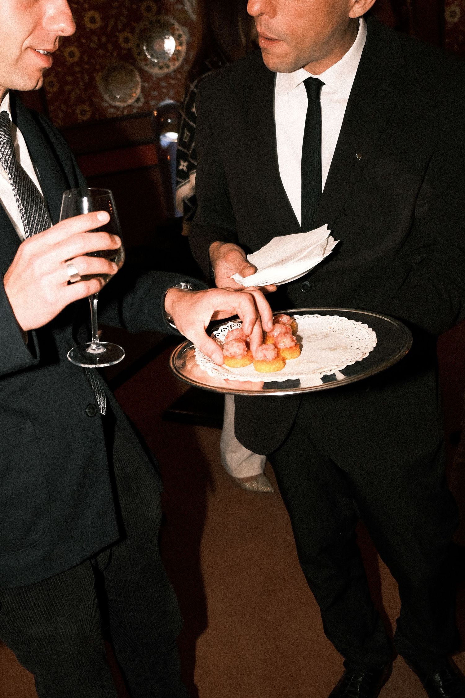 People in formal black suits hold plates and glasses at an elegant cocktail party or reception.