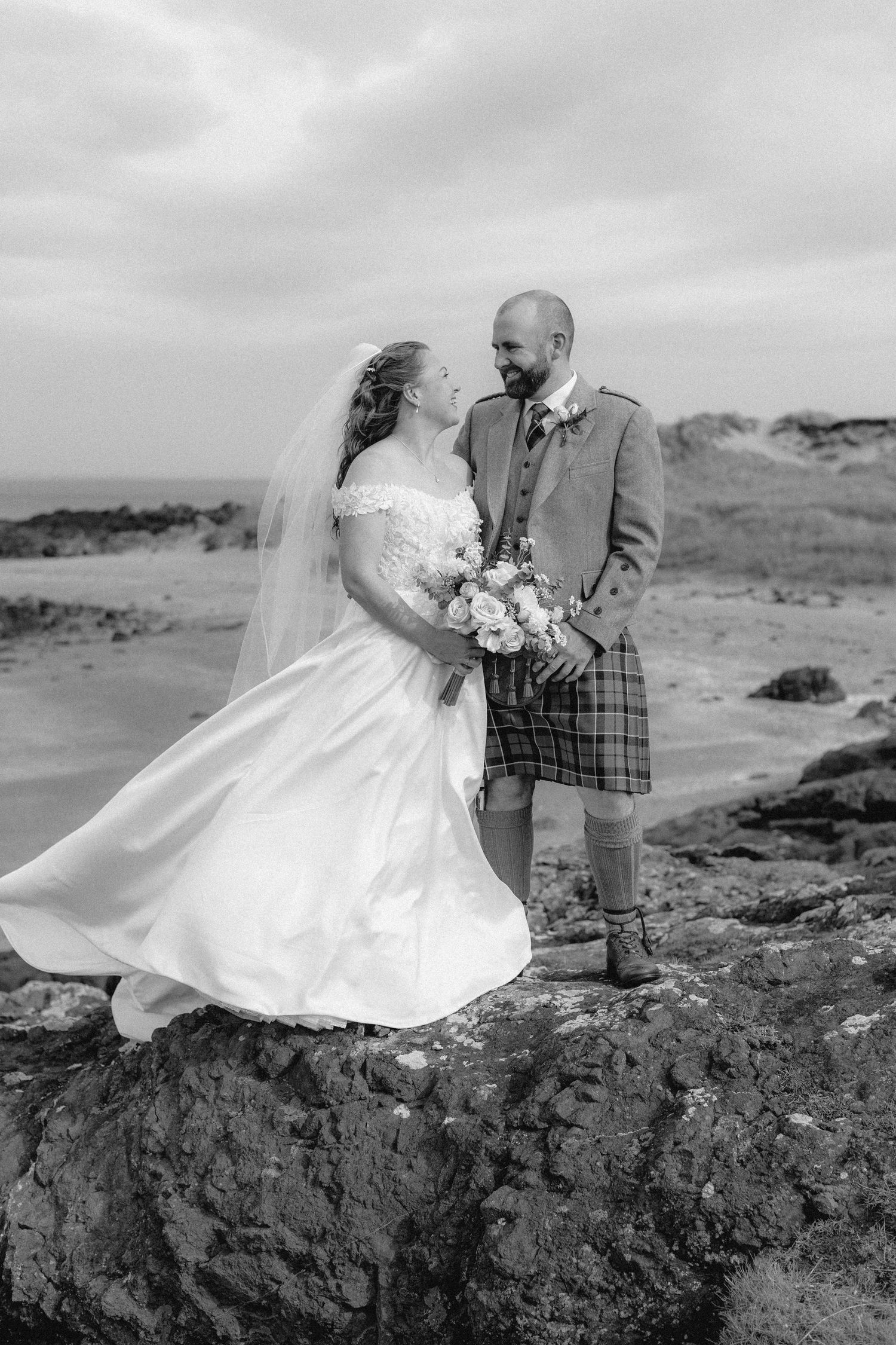 Black and white wedding photo of a couple on rocky coastal cliffs, with the groom wearing a traditional Scottish kilt.