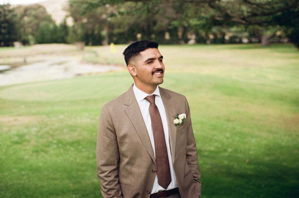 Groom in tan suit with boutonniere smiling outdoors on green golf course lawn at wedding.
