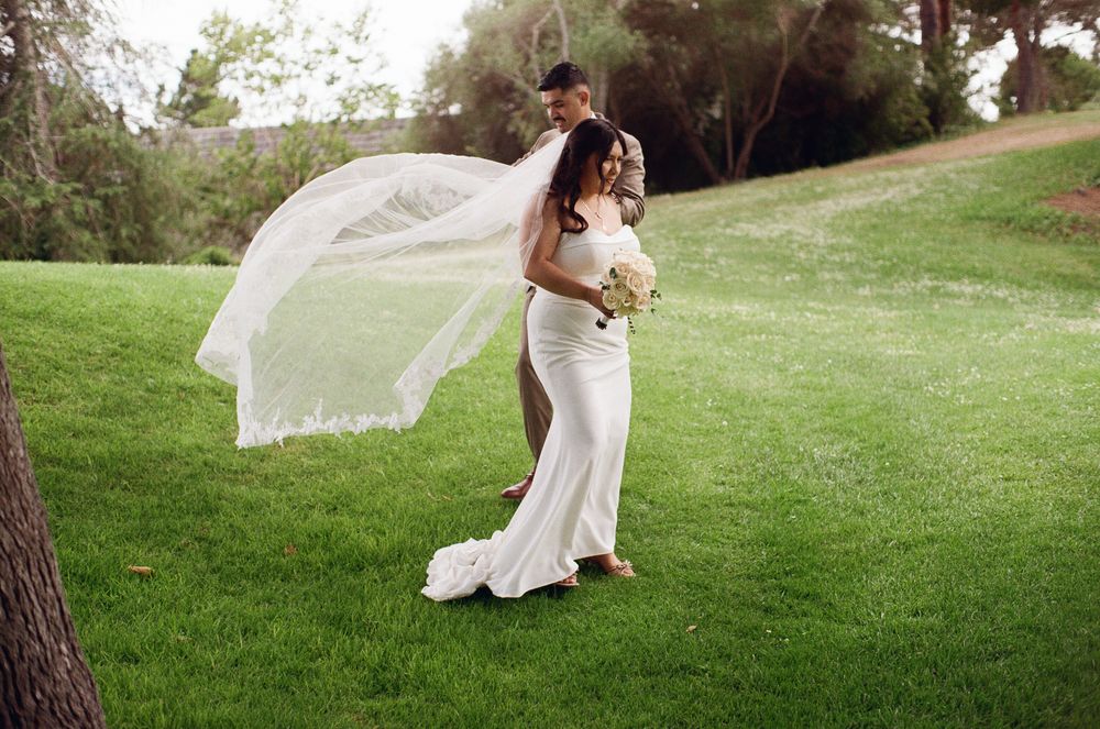 Bride in white gown with flowing veil embraces groom on lush green lawn at outdoor wedding ceremony.
