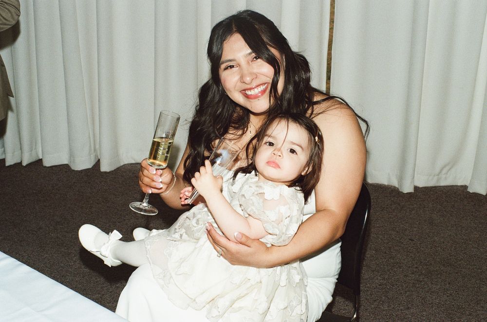 A smiling woman holds a baby in a white dress while raising a champagne glass at a formal event.