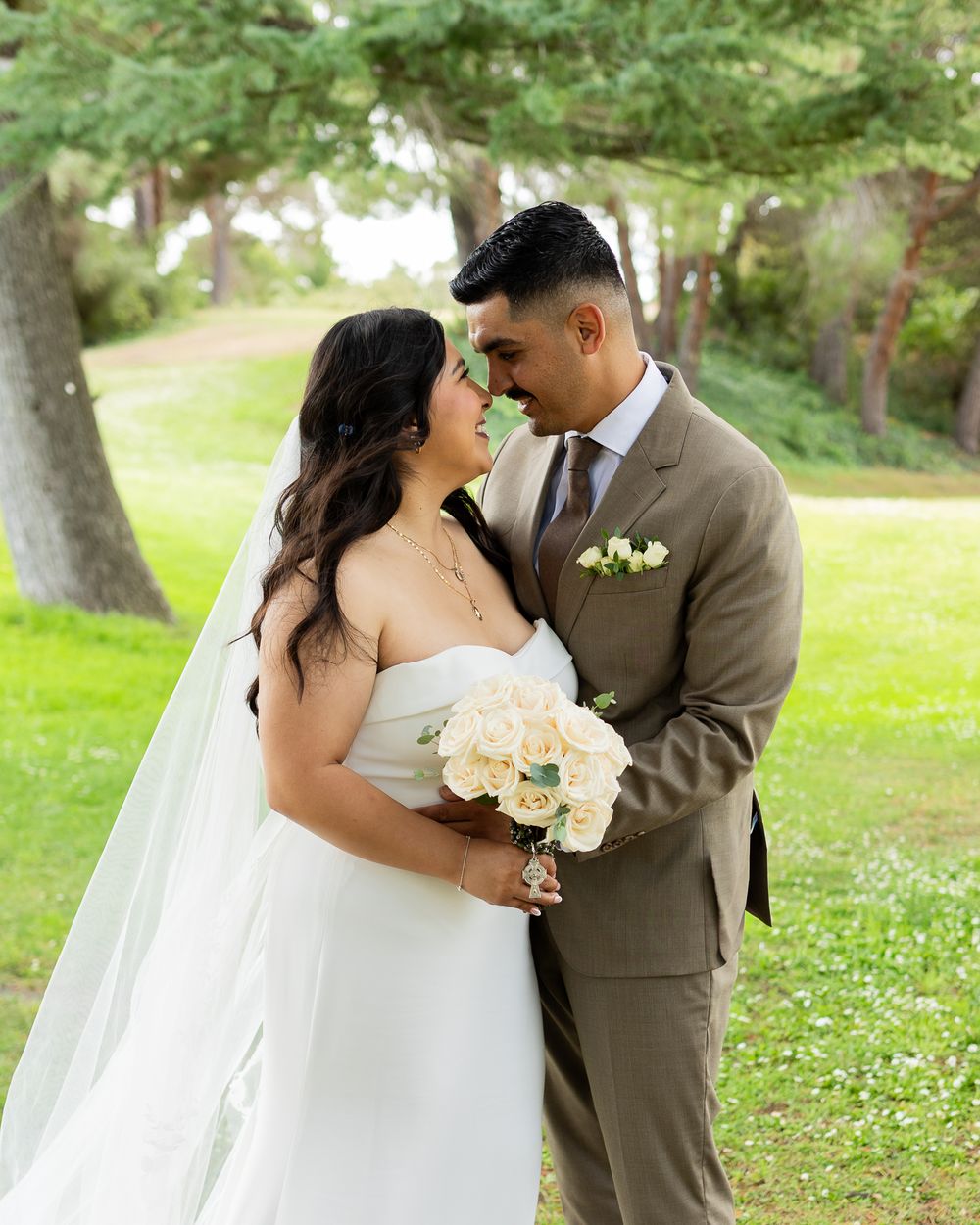 Bride and groom share a romantic moment outdoors, she in a white gown with veil, he in a tan suit, holding white flowers.