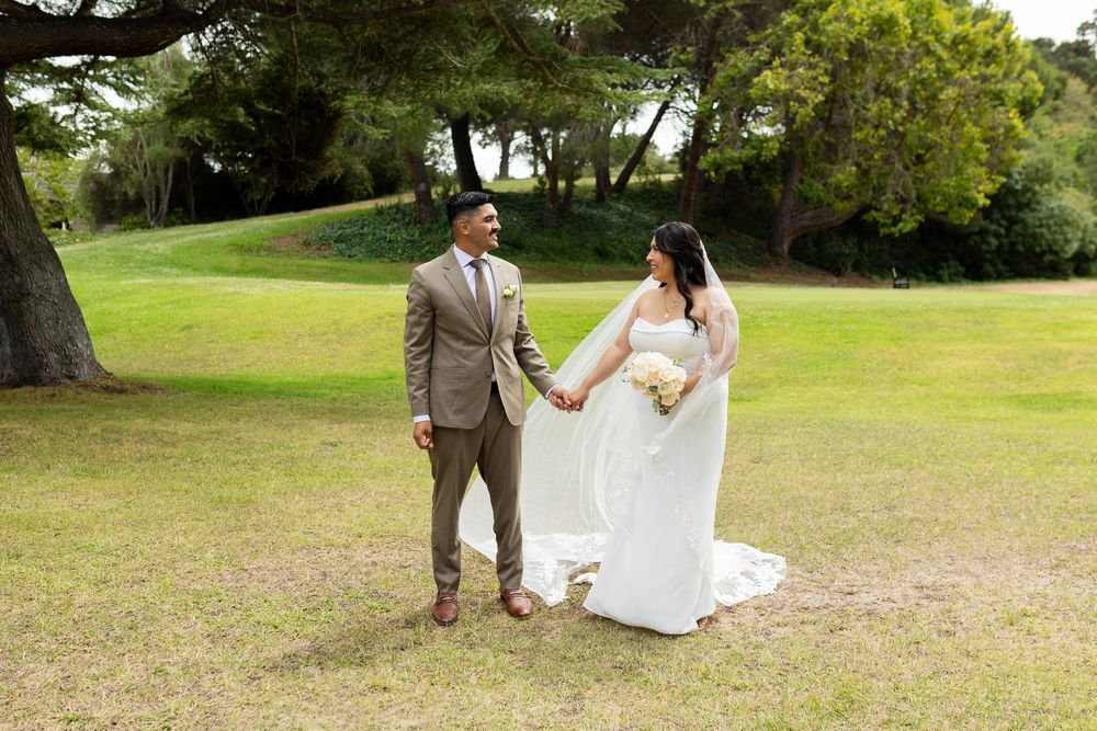 Bride and groom hold hands in a lush green park on their wedding day, bride in white gown with veil.