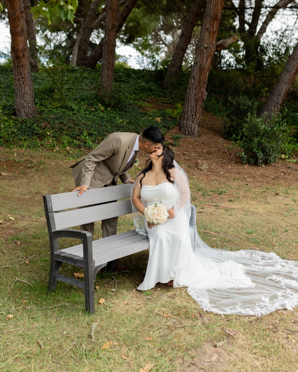Bride in white gown with lace veil sits on bench as groom in tan suit leans in for a kiss outdoors.
