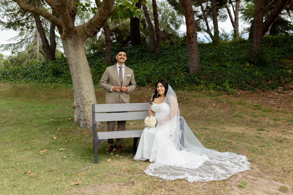 Bride and groom pose outdoors by bench under trees in wedding attire.