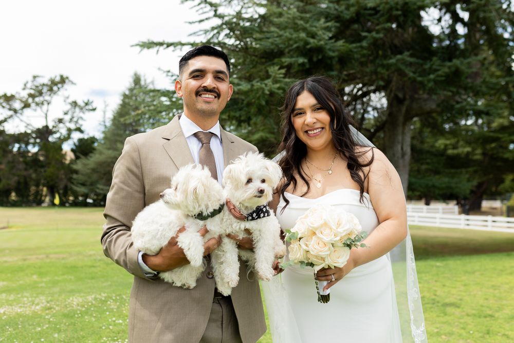 Smiling bride and groom hold a white fluffy dog while posing outdoors on their wedding day.