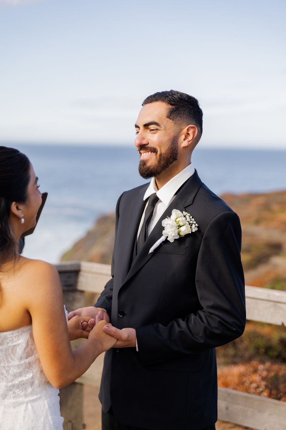 Smiling groom in black suit holding hands with bride outdoors near ocean during wedding ceremony.