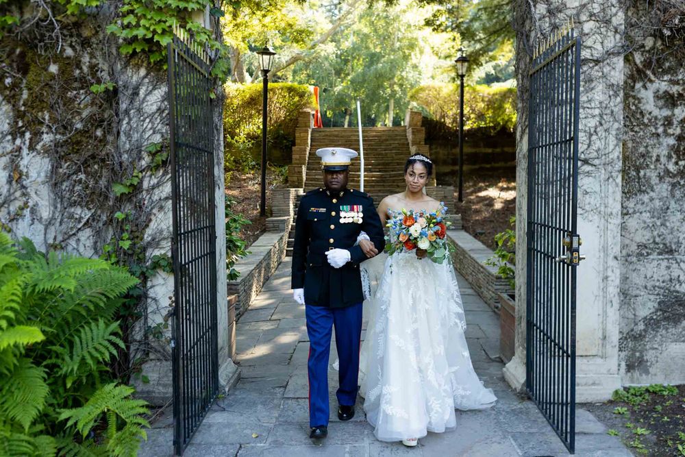 A wedding couple walks through ornate stone gates with lush greenery and ferns along a garden path.