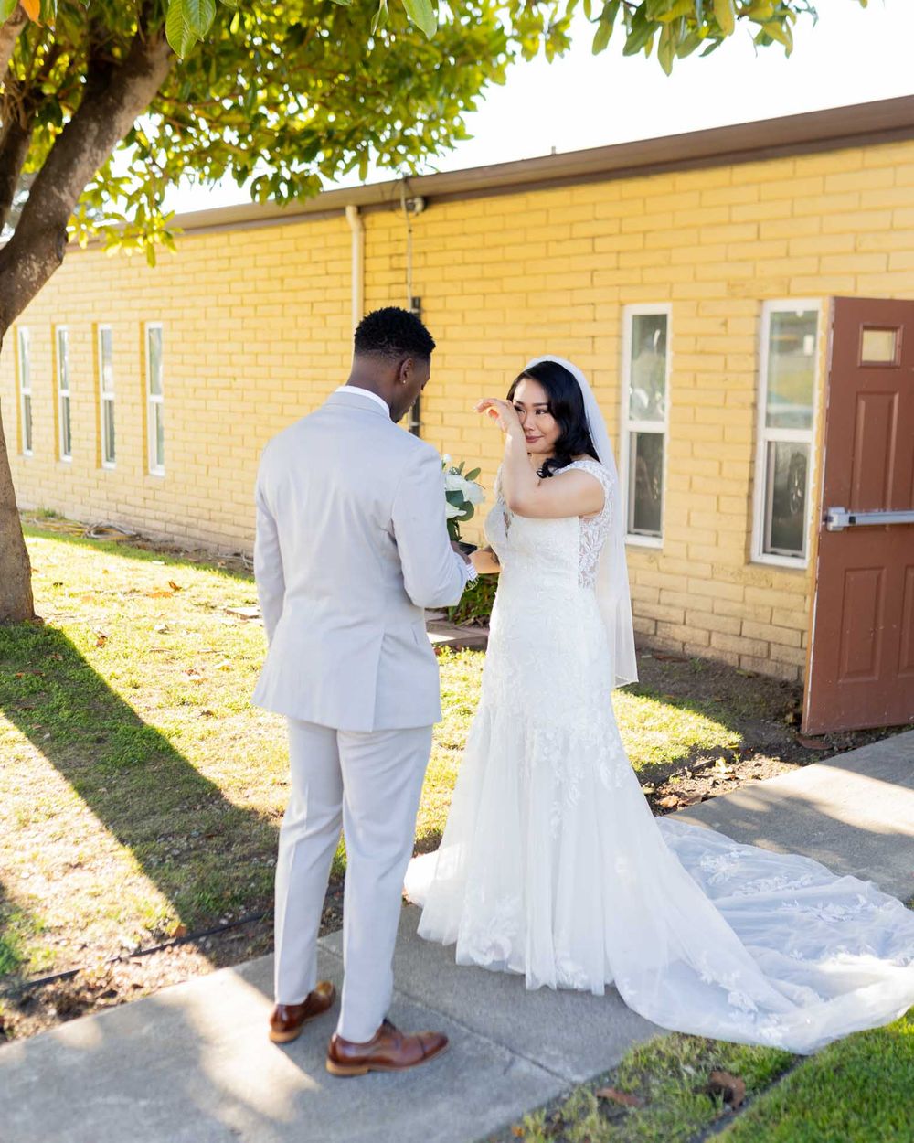 Newlyweds in white attire walk along a beige building exterior during their wedding photoshoot.