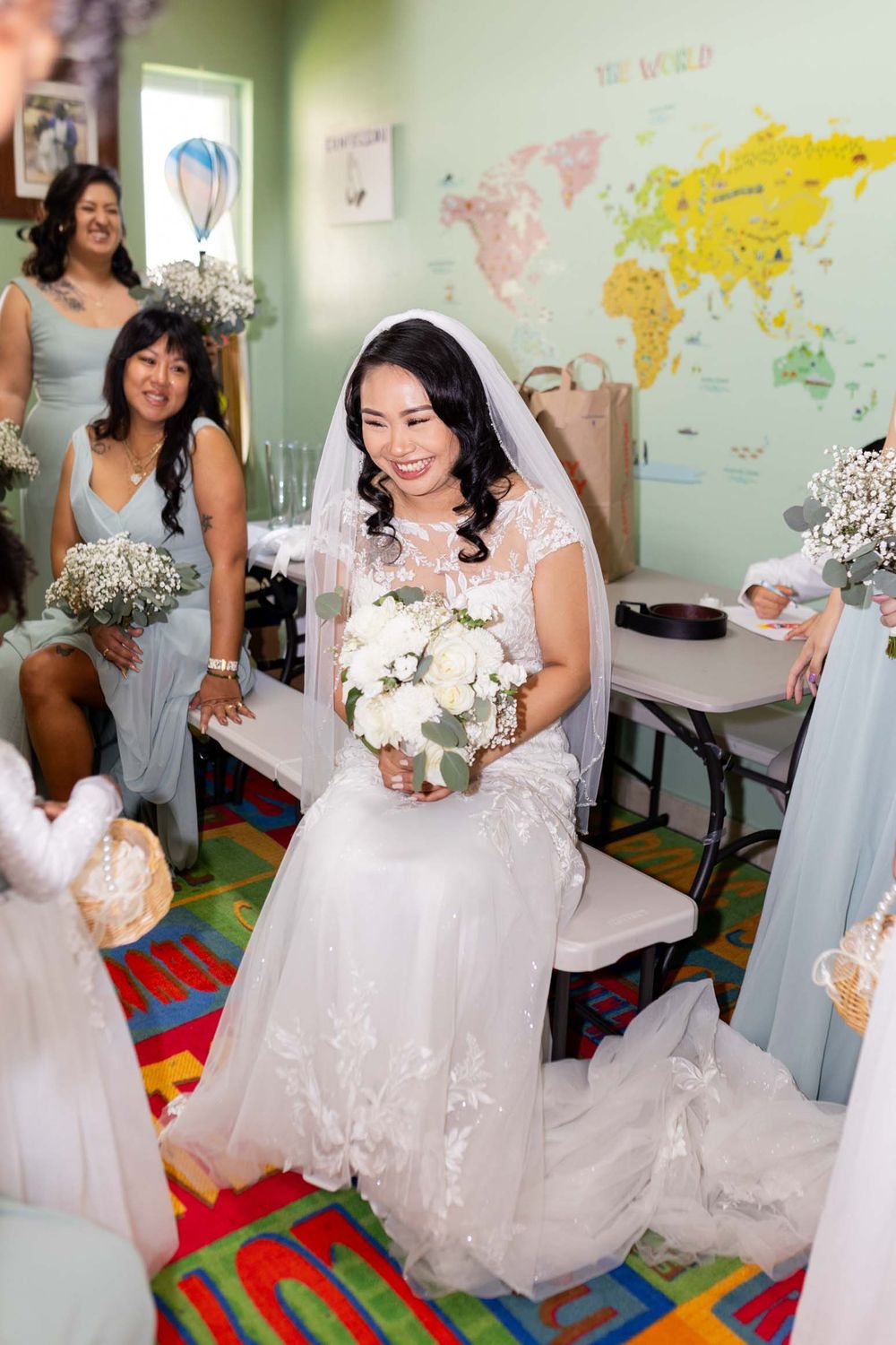 Bridal party members in light blue dresses gather with the bride in a room decorated with world maps on mint green walls.