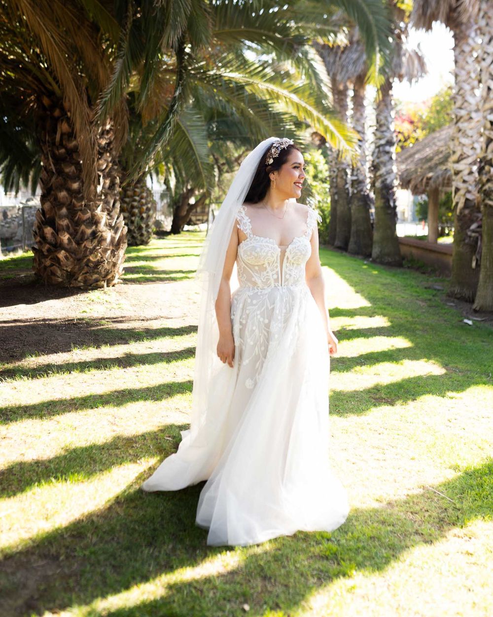 A bride in a flowing white lace dress walks gracefully through a palm tree-lined garden path in golden sunlight.