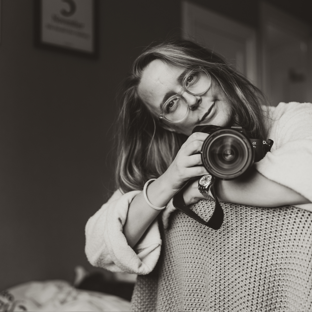Black and white portrait of a photographer holding a professional camera while tilting their head to one side.