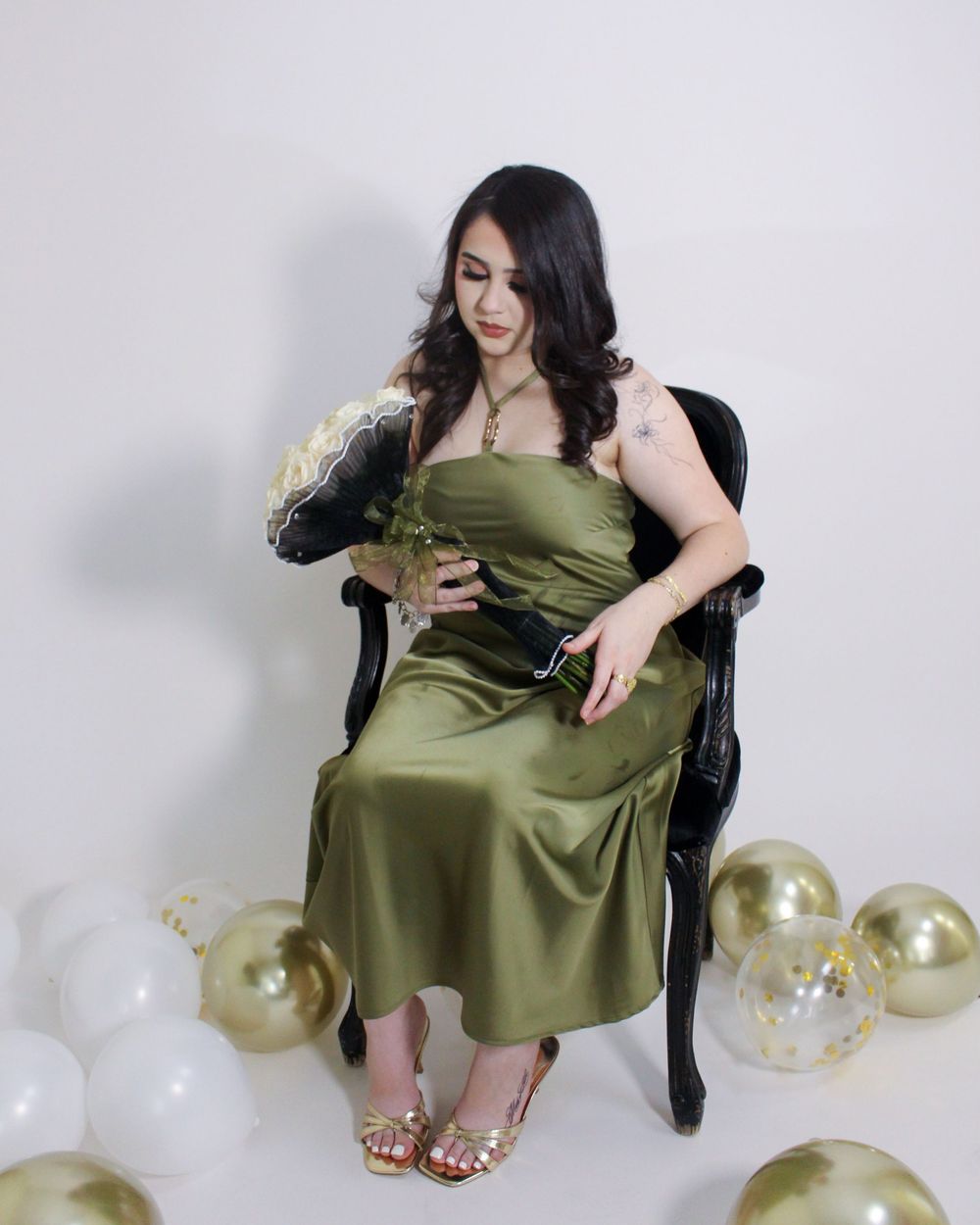 Woman in olive green satin dress seated on black chair surrounded by gold and white balloons.