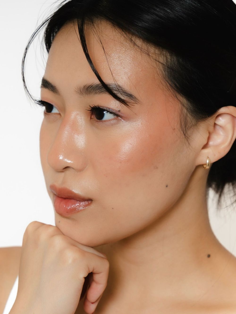 Young woman with glowing skin, subtle blush makeup, gold hoop earring, and dark hair pulled back, posing against white background.