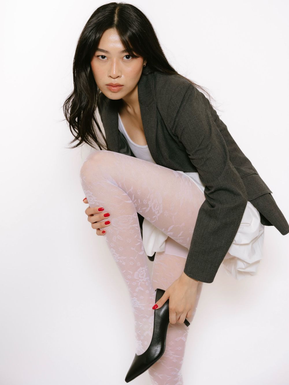 Woman in gray blazer, white skirt, pink lace tights, and black heels poses against white background.
