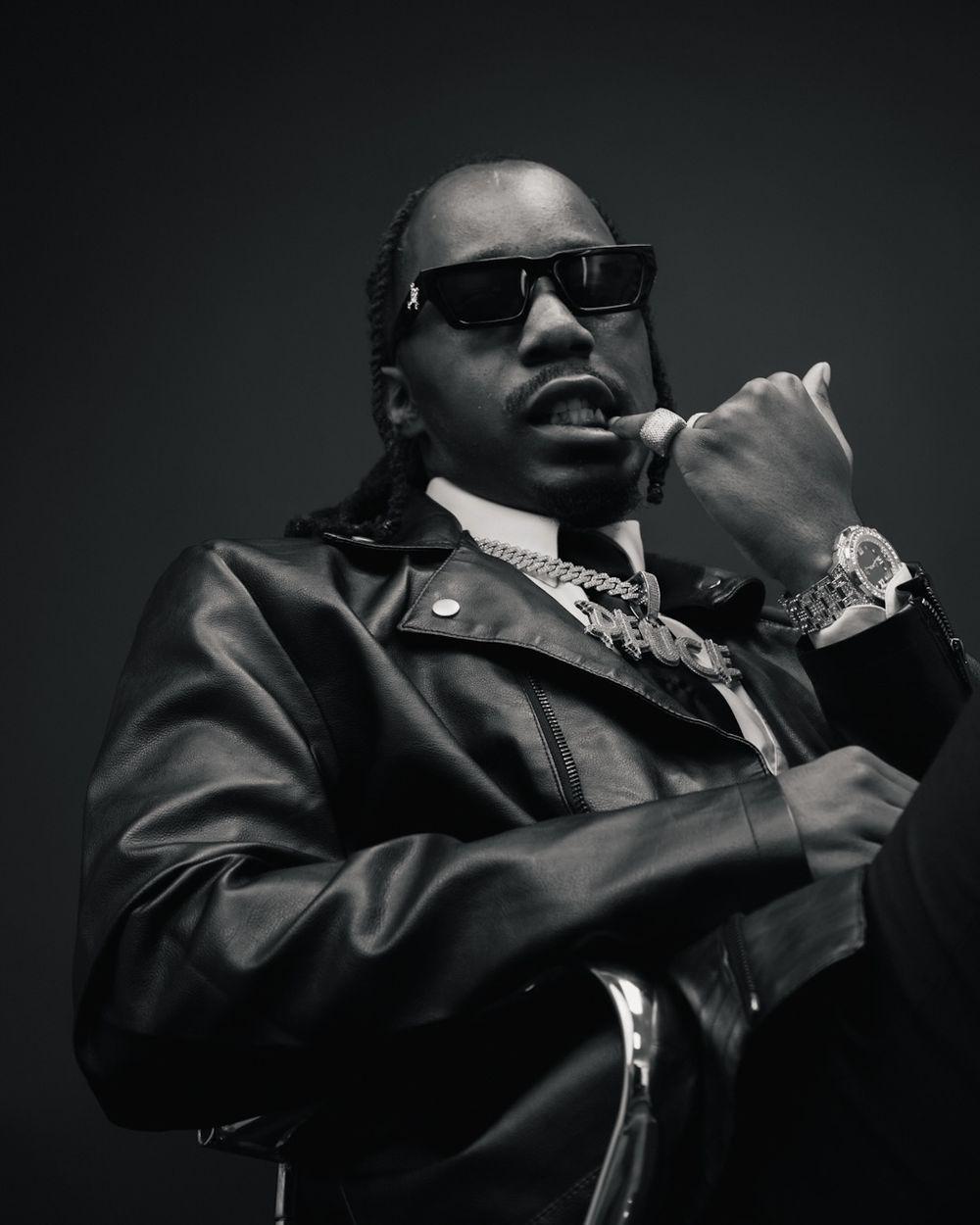 Black and white portrait of a man in leather jacket wearing sunglasses and diamond chain jewelry posing confidently.