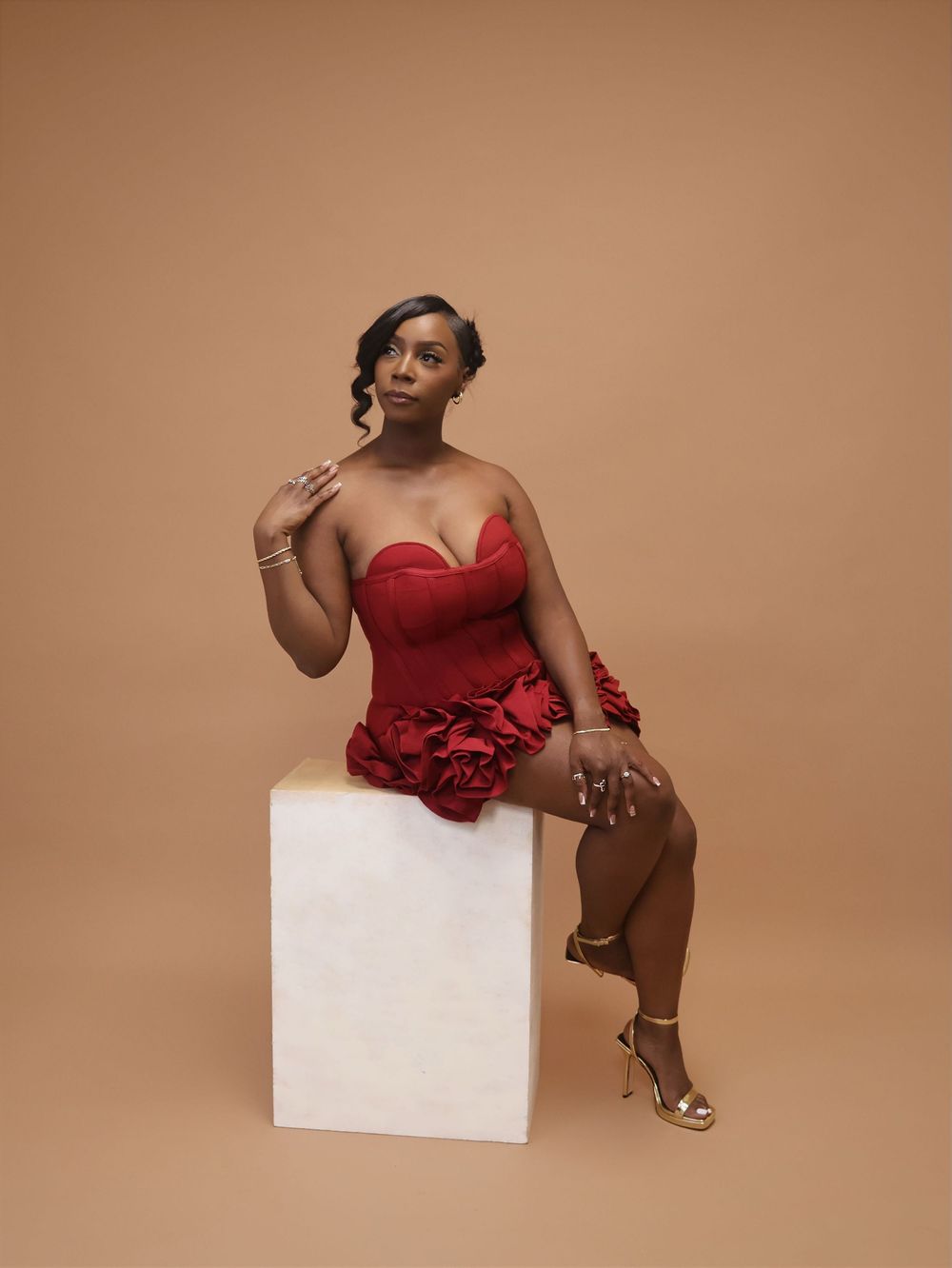 A woman in a red strapless ruffled mini dress and gold heels poses seated on a white cube against a tan background.