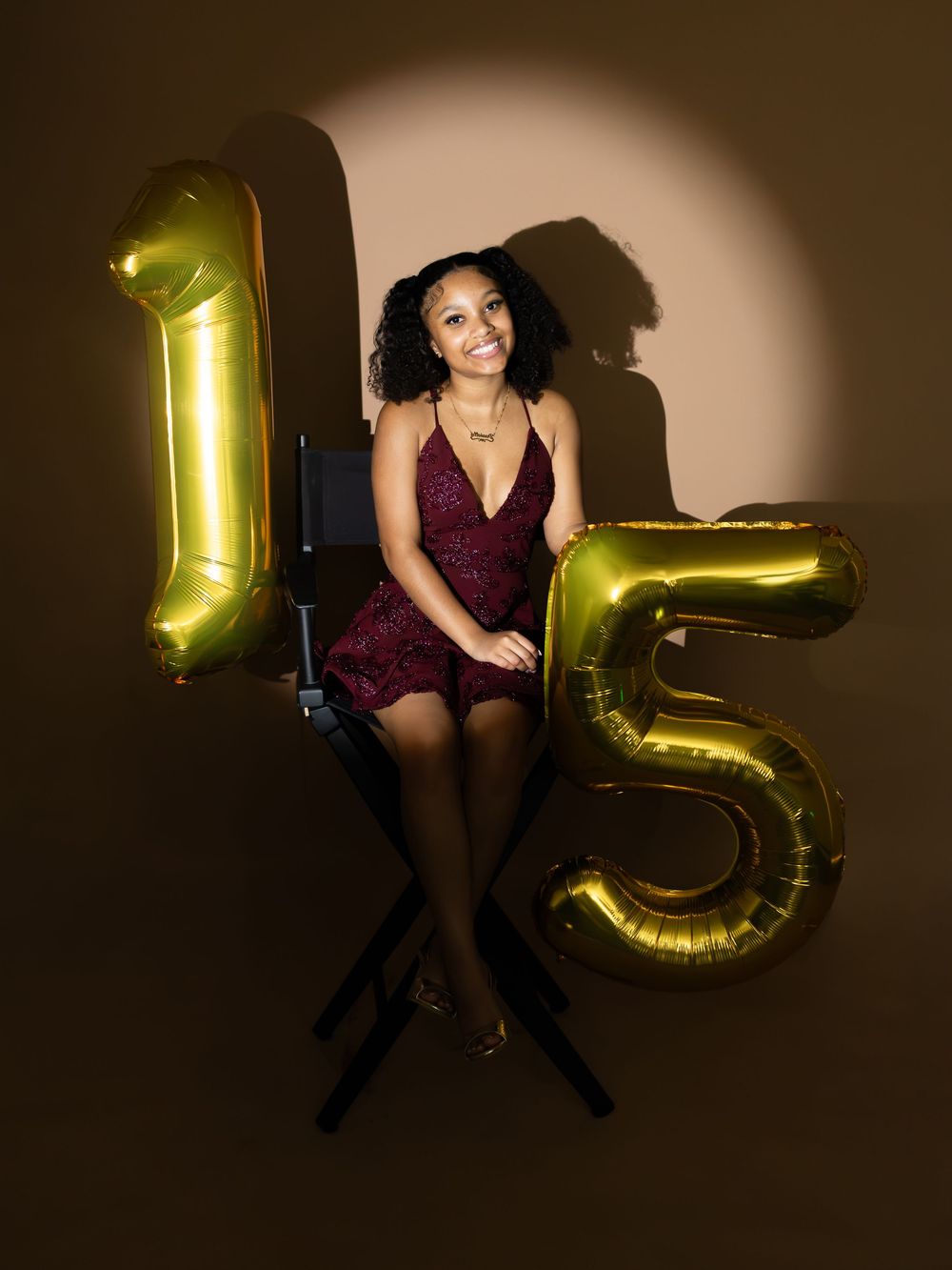 Young woman in burgundy dress poses between gold number 15 balloons for a quinceañera celebration photo.