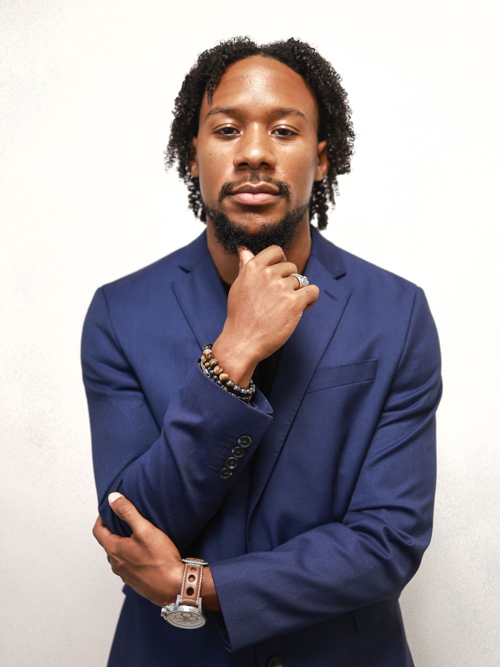 A confident man in a navy blue suit poses thoughtfully with hand on chin, wearing a watch and bracelets.