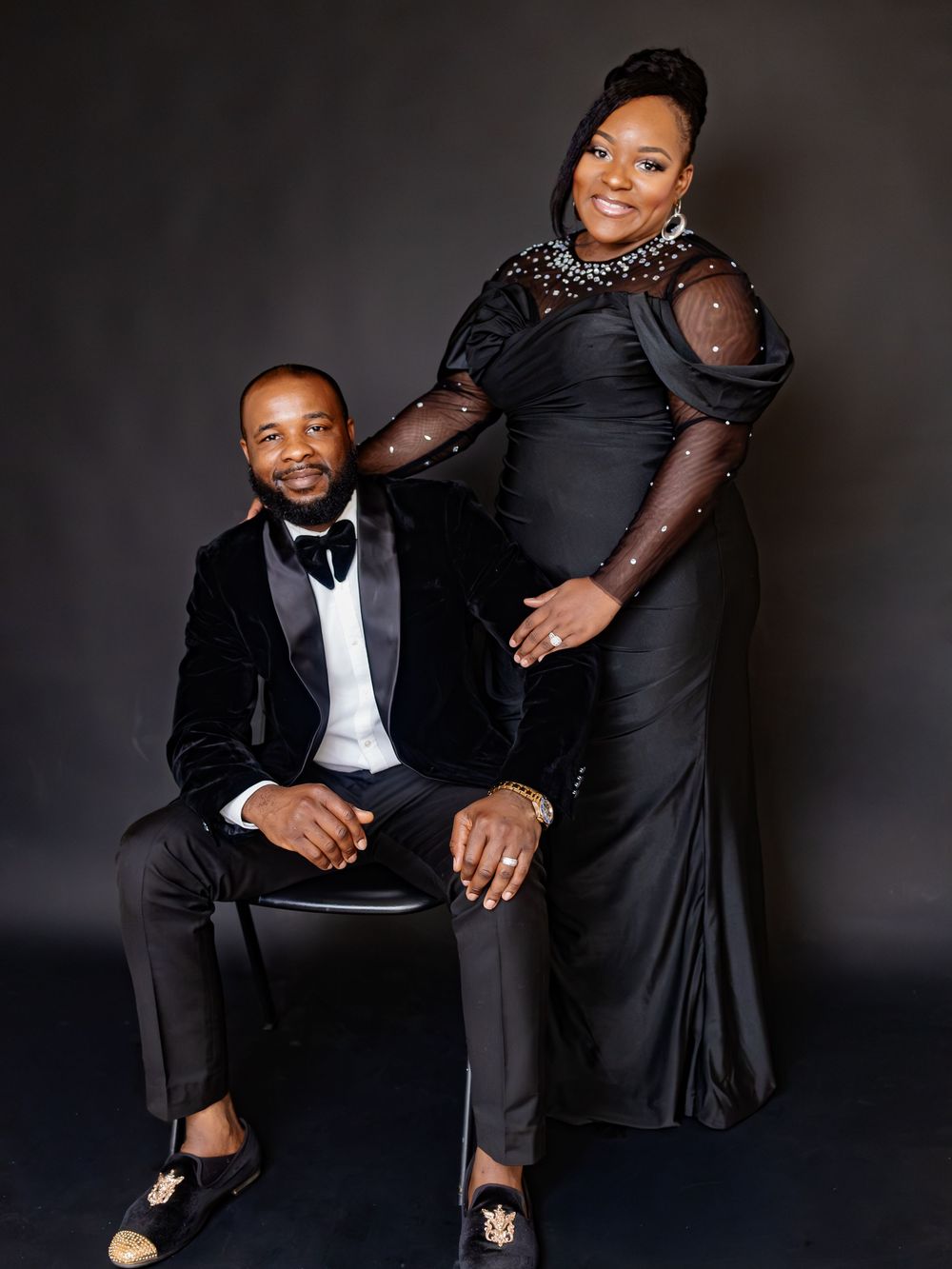 Elegant couple in matching black formal attire posing for a professional portrait against a dark studio background.