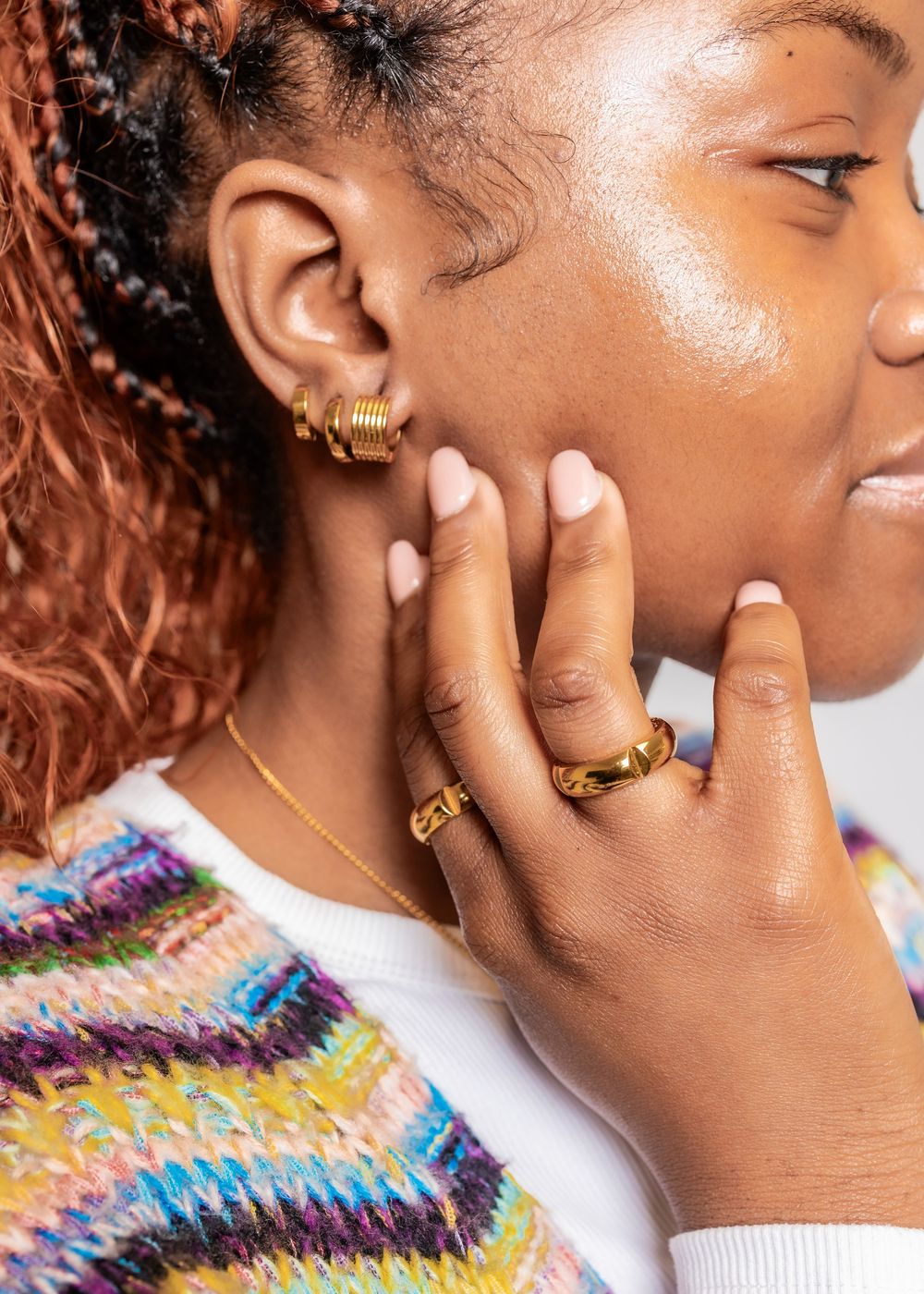 A person wearing a colorful knit sweater and gold jewelry touches their face while looking to the side in profile.