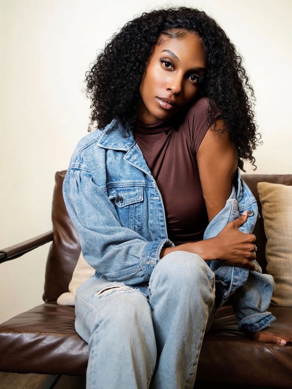 A young woman with curly hair wearing a denim jacket and brown top sits on a leather couch posing.