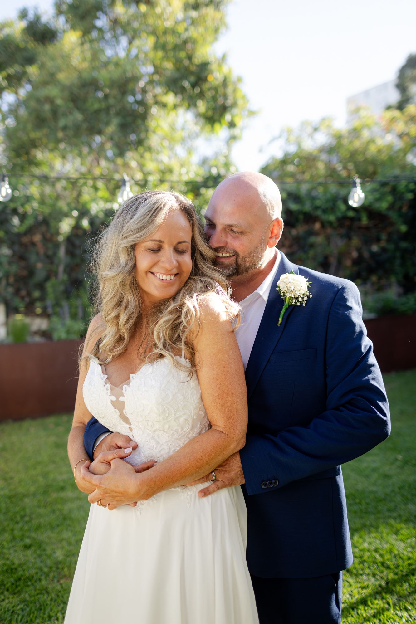 A couple embraces outdoors in wedding attire, with the bride in white lace and groom in navy suit on a sunny garden day.
