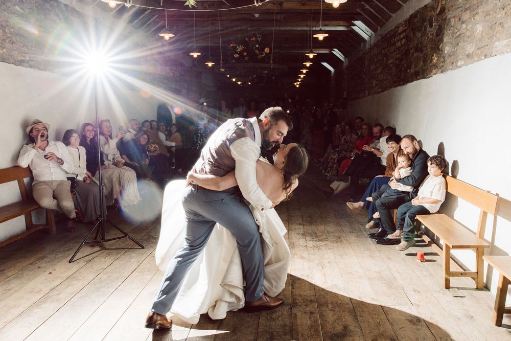A couple shares their first dance at a rustic barn wedding venue with guests watching from wooden benches.