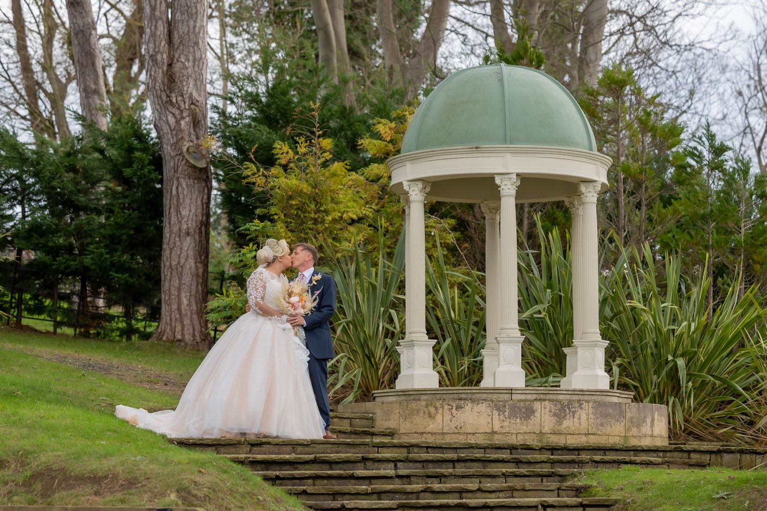 A couple stands together near an elegant stone gazebo with a domed roof surrounded by lush garden landscaping. Rushpool Hall Wedding