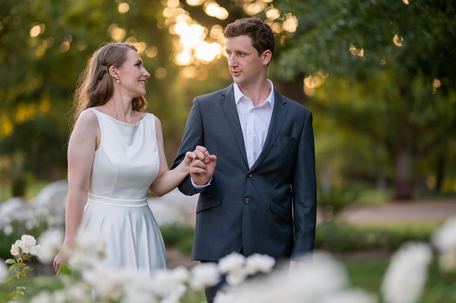 Couple holding hands in a garden at sunset, woman in white dress and man in gray suit with white roses.