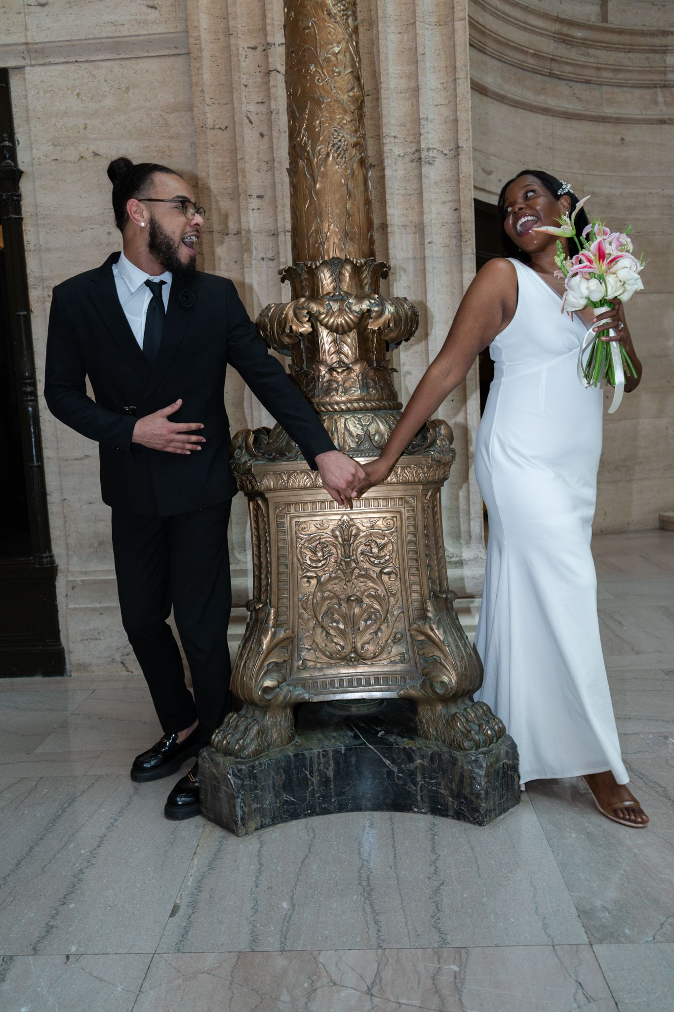 Interracial couple holds hands laughing beside ornate gold column in marble wedding venue.