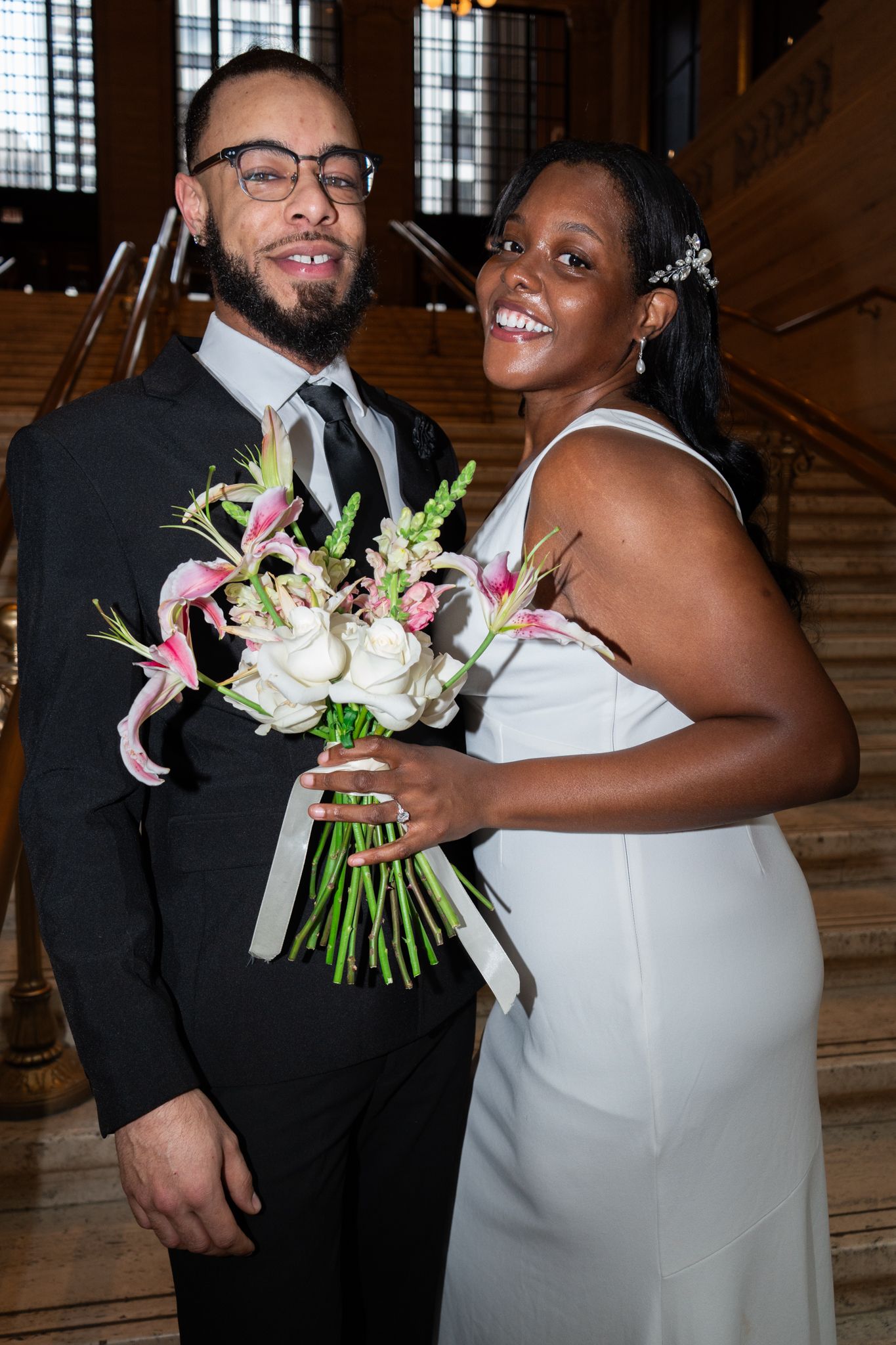 Smiling interracial couple poses together on grand staircase, bride holding pink lily and white rose bouquet.