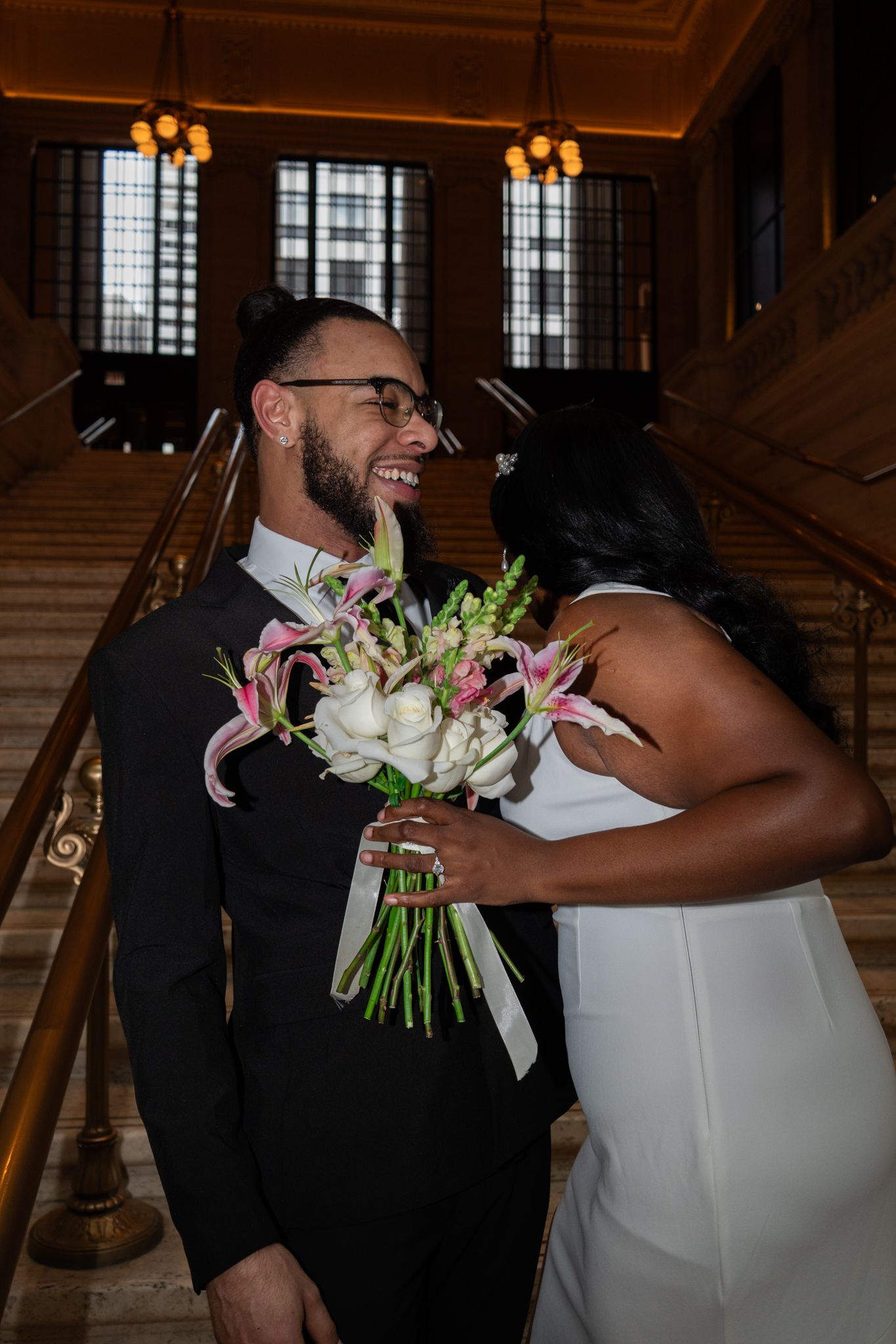 Groom in black suit laughs joyfully as bride in white dress presents pink and white wedding bouquet.