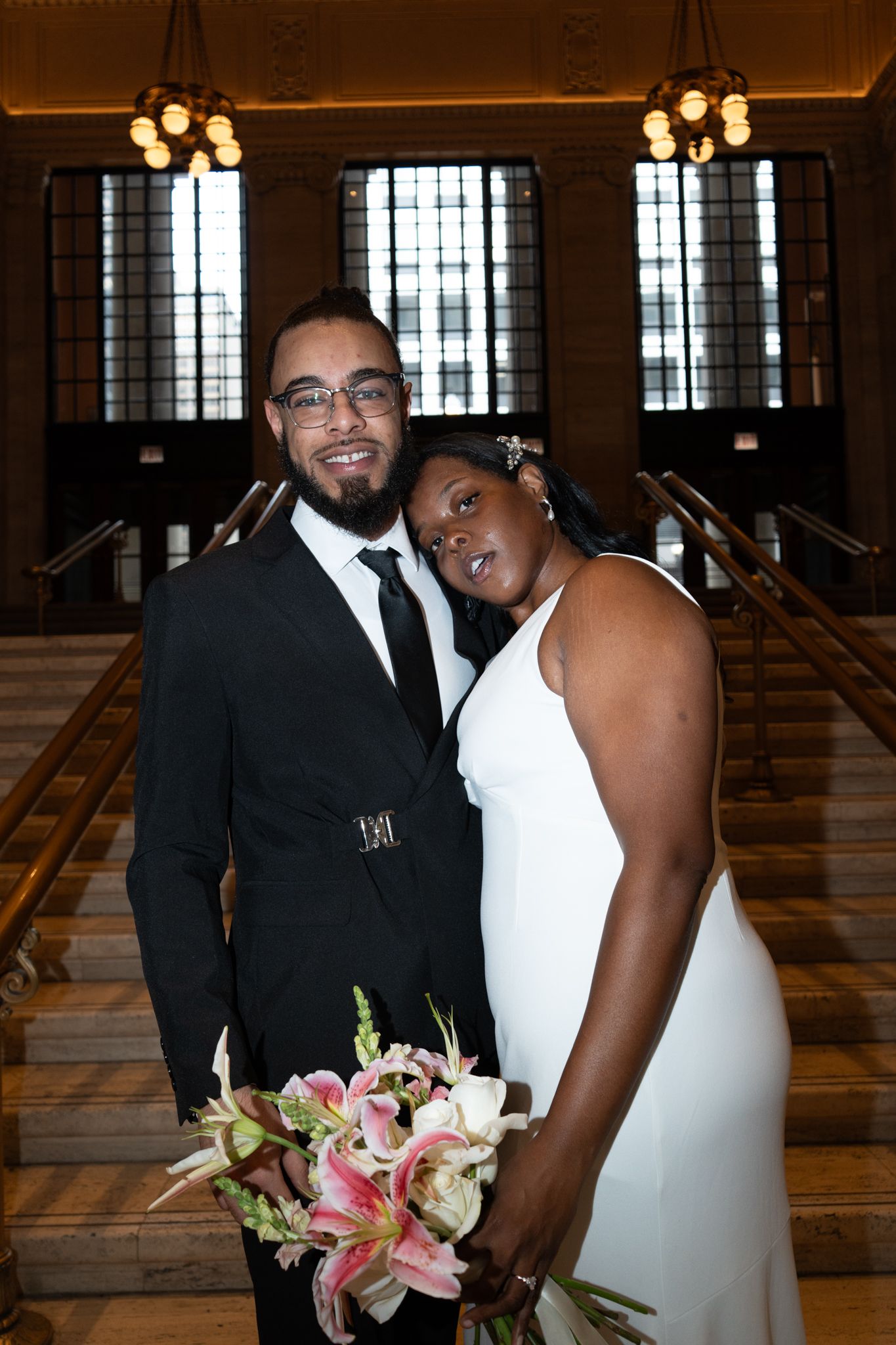 A couple poses in wedding attire inside an elegant hall, the bride holding pink lilies bouquet.