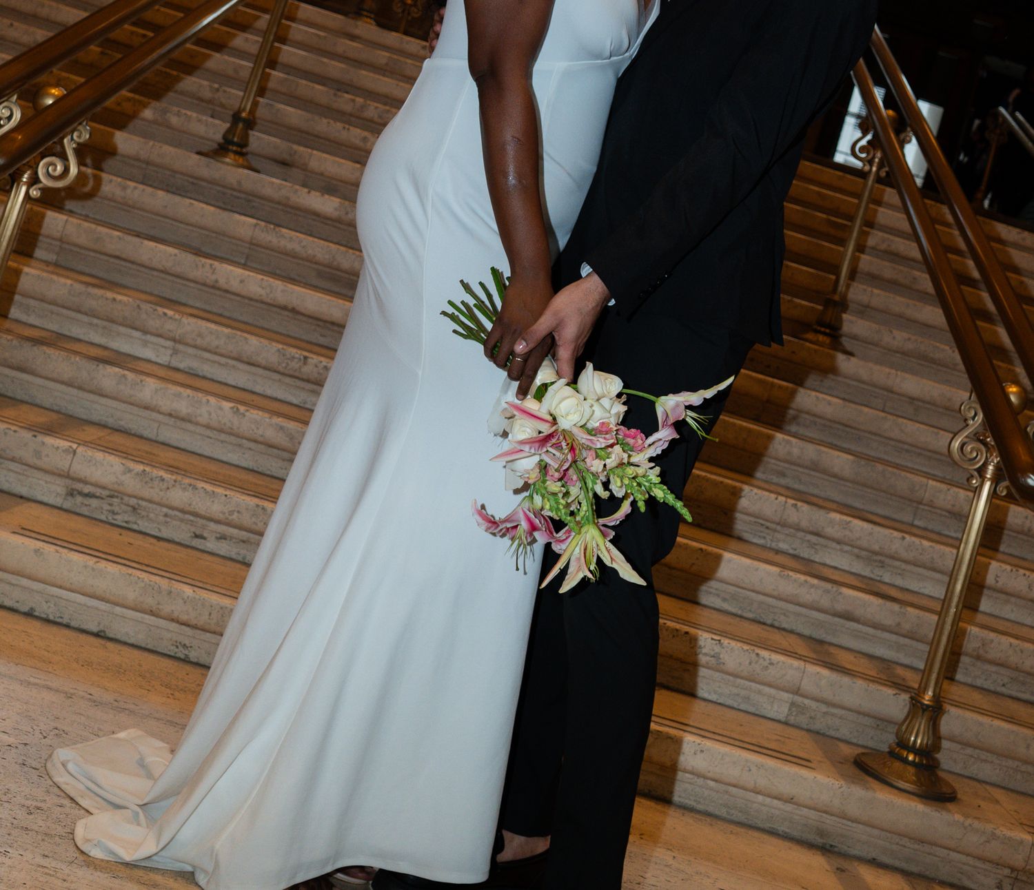 Bride in white gown and groom hold hands with floral bouquet on marble staircase.