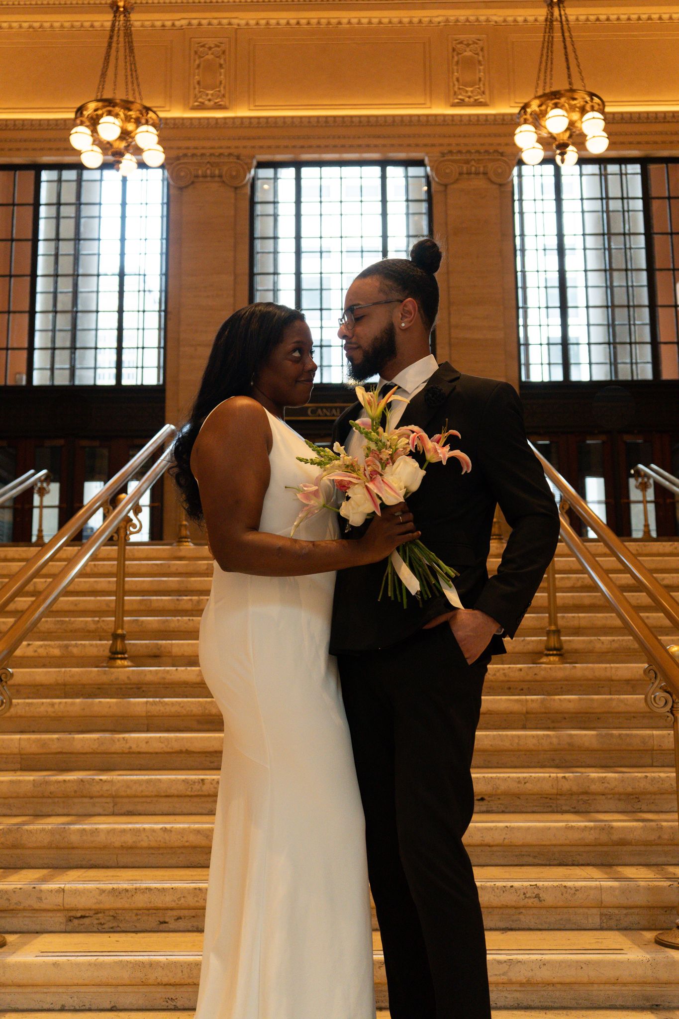Bride and groom in wedding attire share a romantic moment on grand staircase holding a pink and white bouquet.