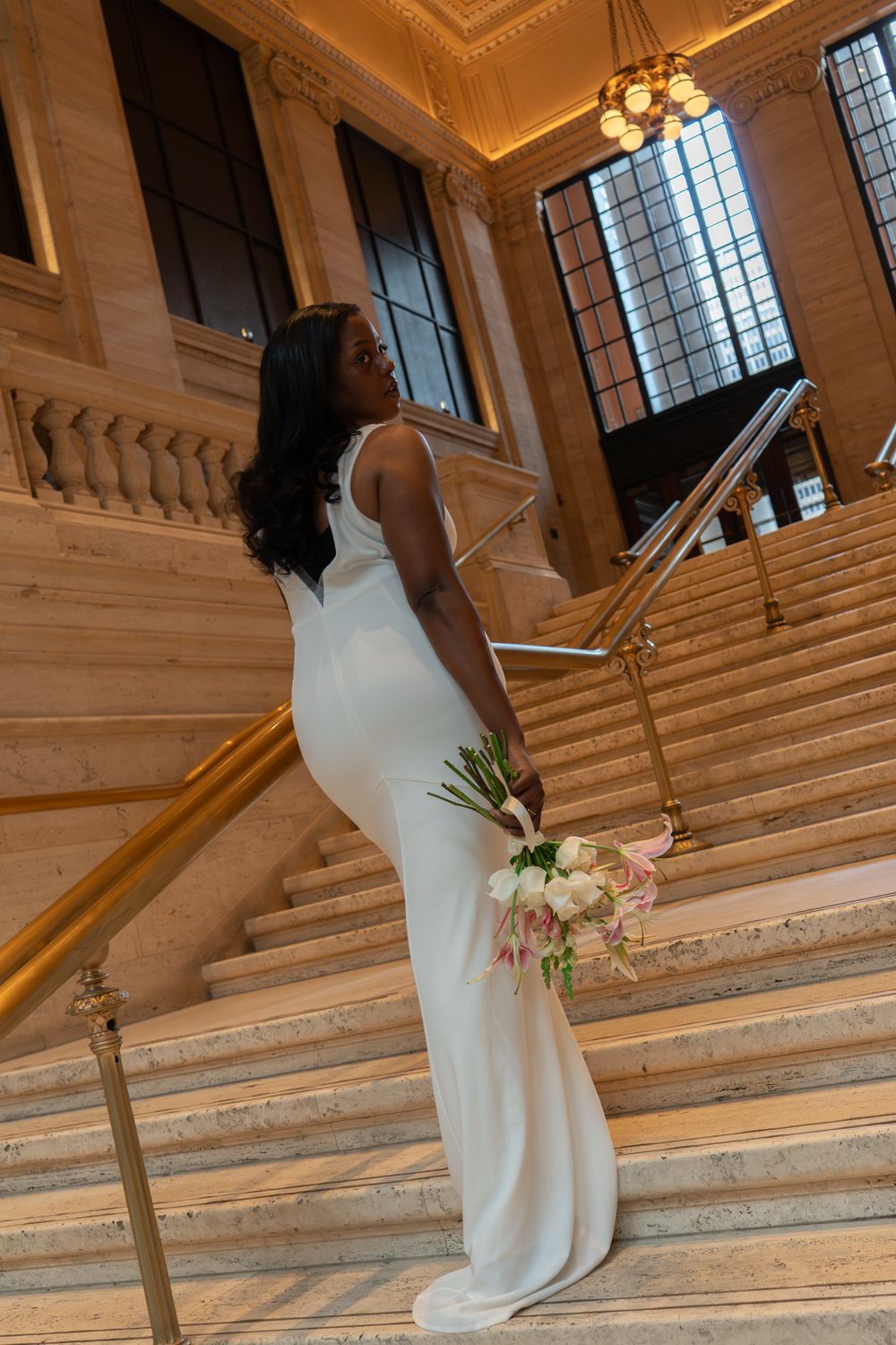 Elegant bride in white gown holds bouquet while posing on ornate marble staircase in grand building.