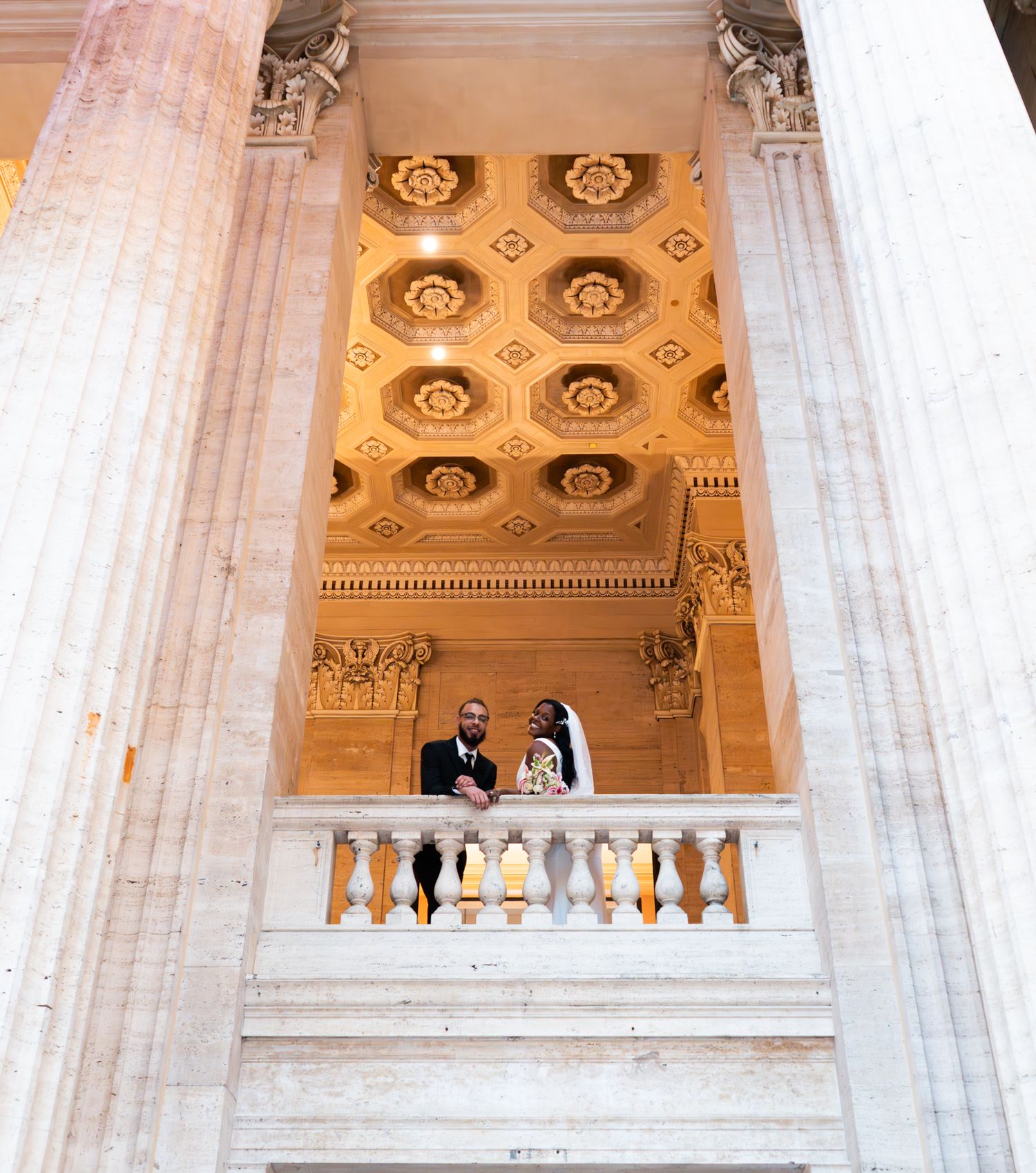 Bride and groom pose on ornate balcony of grand marble building with gilded coffered ceiling.